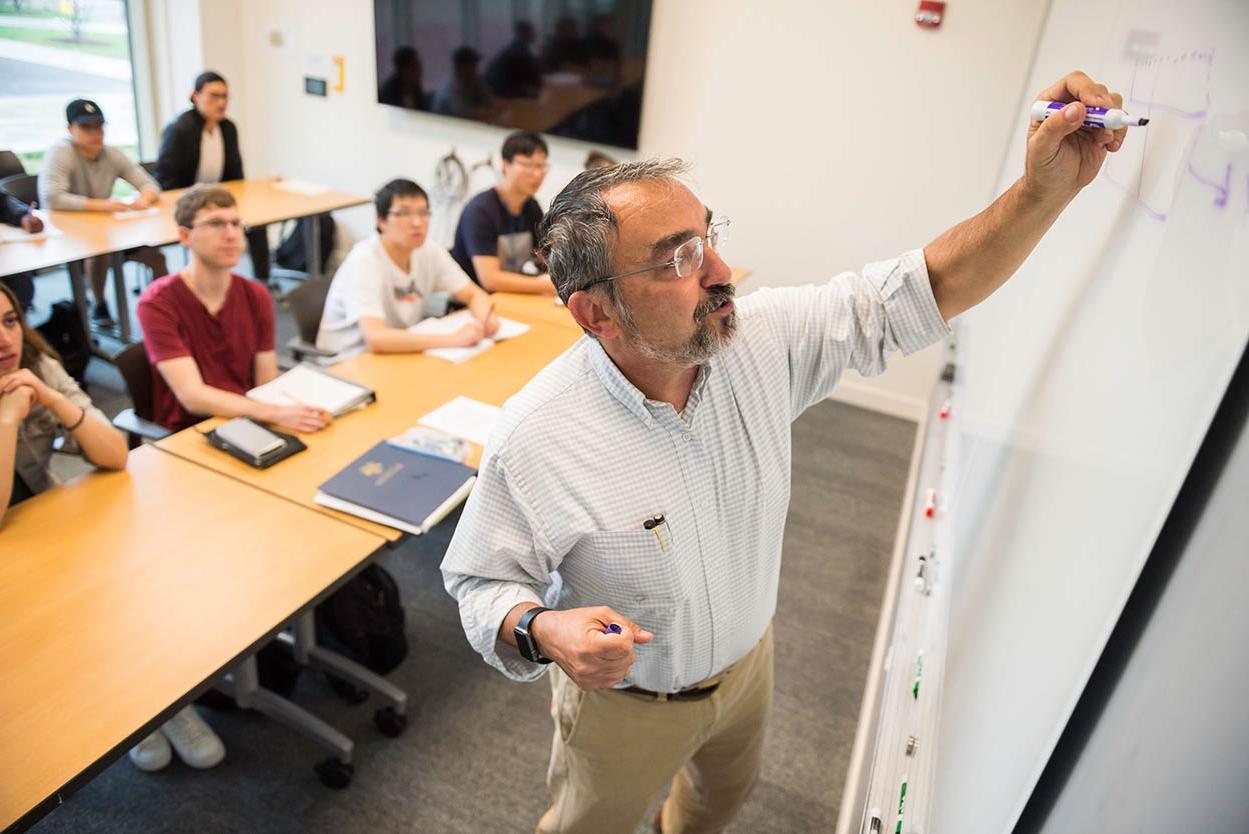 Professor writing on a whiteboard at the front of a classroom